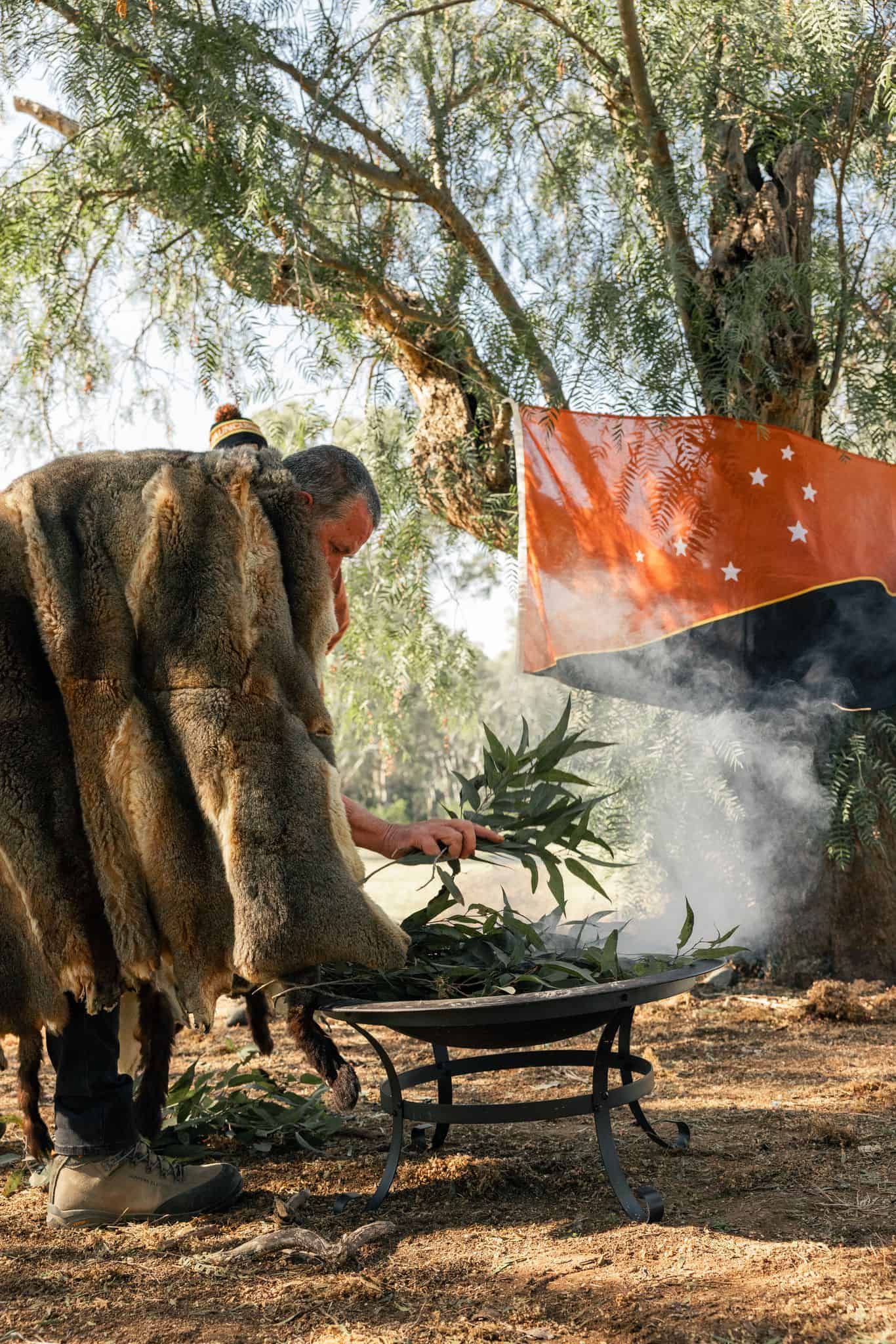 Elder lights the ceremonial fire, with possum skin cloak on and Taungurung flag in the background