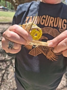 Person weaving a small yellow straw flower with fingers, wearing a dark T‑shirt and rings on one hand (craft activity)