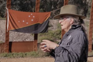 Side profile of an elderly woman wearing a wide beige hat and green-rimmed glasses, gesturing outdoors near an orange banner on a wooden frame