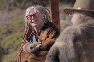 Smiling elderly woman with red glasses and gray hair in a fur coat outdoors, talking to someone wearing a wide-brim hat.
